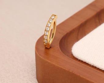 May include: A gold-colored curved earring with a row of clear, sparkling stones. The earring is set against a wooden surface with a white fabric insert, creating a contrast of textures and colors. The design is simple yet elegant.