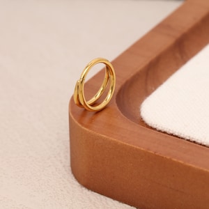 May include: A gold-colored, double-hoop ring. The ring is made of a shiny, metallic material. The ring is resting on a wooden tray with a white fabric insert.