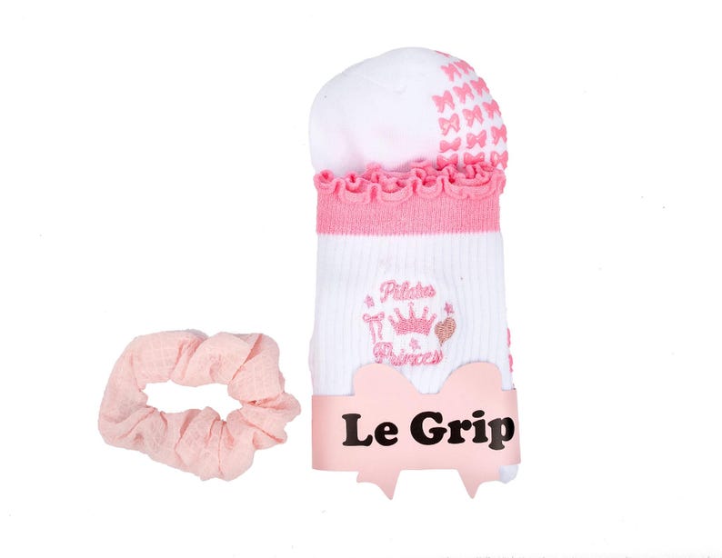 May include: White and pink Pilates socks with a ruffled cuff and bow accents, paired with a matching pink scrunchie. The socks feature the text "Pilates Princess" and the brand name "Le Grip".