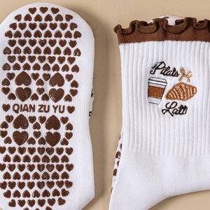 May include: White socks with brown heart-shaped grips on the soles. The top of the socks features a brown ruffled trim and an embroidered design of a coffee cup, croissant, and the words "Pilate Latte". The text "QIAN ZU YU" is printed on the sole.