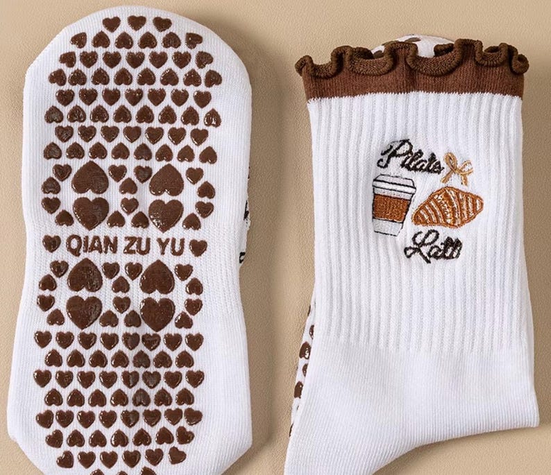 May include: White socks with brown heart-shaped grips on the soles. The top of the socks features a brown ruffled trim and an embroidered design of a coffee cup, croissant, and the words "Pilate Latte". The text "QIAN ZU YU" is printed on the sole.