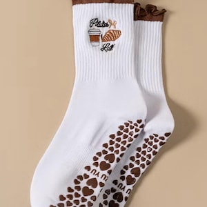 May include: White socks with brown accents and a heart pattern on the soles. The socks feature the embroidered text "Pilates Latte" with a coffee cup and a pastry. The top edge has a ruffled brown trim.