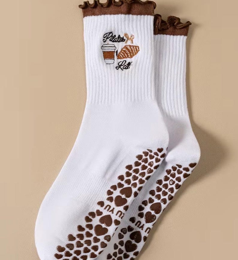 May include: White socks with brown accents and a heart pattern on the soles. The socks feature the embroidered text "Pilates Latte" with a coffee cup and a pastry. The top edge has a ruffled brown trim.