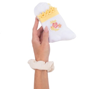Op de afbeelding: Witte babysok met gele ruches en een geel en roze bloemenmotief. De sok heeft de tekst "Island Time" geborduurd. Een crèmekleurige scrunchie zit om de pols.