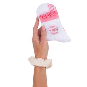 May include: White Pilates socks with pink accents, including a ruffled trim and bow details. The socks feature the text "Pilates a Princess" with a crown and flamingo illustration. The socks are held up against a white background.