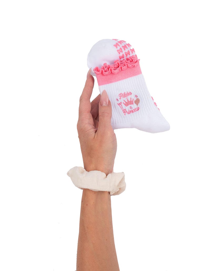May include: White Pilates socks with pink accents, including a ruffled trim and bow details. The socks feature the text "Pilates a Princess" with a crown and flamingo illustration. The socks are held up against a white background.