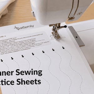 May include: A white sewing machine with a practice sheet that says "Beginner Sewing Practice Sheets." The sheet has curved lines for practicing stitches. A spool of thread is visible on the left side of the image.