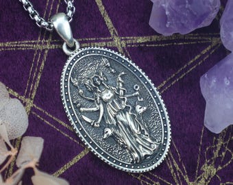 May include: A silver-toned pendant necklace featuring an oval pendant with a detailed relief of a goddess figure. The pendant hangs from a chain. The background is a deep purple fabric with gold accents, and amethyst crystals.