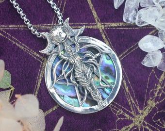 May include: A silver pendant necklace featuring a detailed figure within a circular frame. The pendant has an abalone shell inlay and is suspended from a silver chain. The background is a deep purple fabric with gold accents, and clear crystals are scattered around.