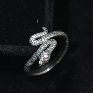 May include: A silver-toned metal ring shaped like a coiled snake with textured scales. The snake's head is embellished with small, clear white stones and a single, tiny orange stone for the eye, creating a detailed and realistic serpent design.
