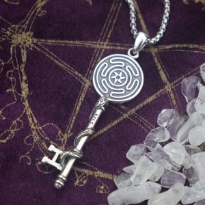 May include: A silver pendant necklace with a key design. The key has a round pendant with a spiral pattern and a star in the centre. The key's handle is detailed with a snake design. The necklace is on a silver chain.
