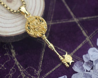 May include: A gold-colored key pendant necklace. The key features a detailed circular head with an ornate design and a long, slender stem. The necklace chain is also gold-colored. The background is a dark purple fabric with gold-colored lines and a wooden slice.