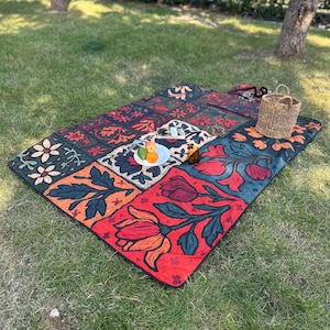 May include: A patterned picnic blanket in shades of red, orange, and green, with a floral design. The blanket is rectangular and placed on grass. A wicker basket and a plate of fruit are on the blanket.