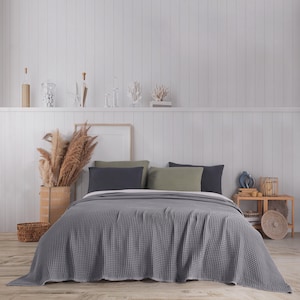 Puede incluir: Una colcha gris con tejido de gofre cubre una cama, complementada con almohadas gris oscuro y verde oliva. La habitación presenta una pared con paneles blancos, una mesita de noche de madera y elementos decorativos, creando un ambiente sereno.