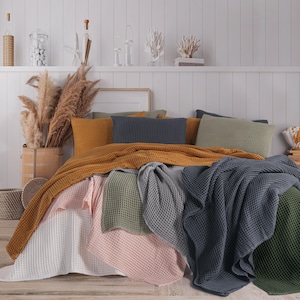 May include: A bed displays a collection of waffle-weave blankets and cushions in mustard, grey, pink, white, and olive green. The blankets are arranged over the bed, creating a layered look. Decorative items are on a shelf in the background.