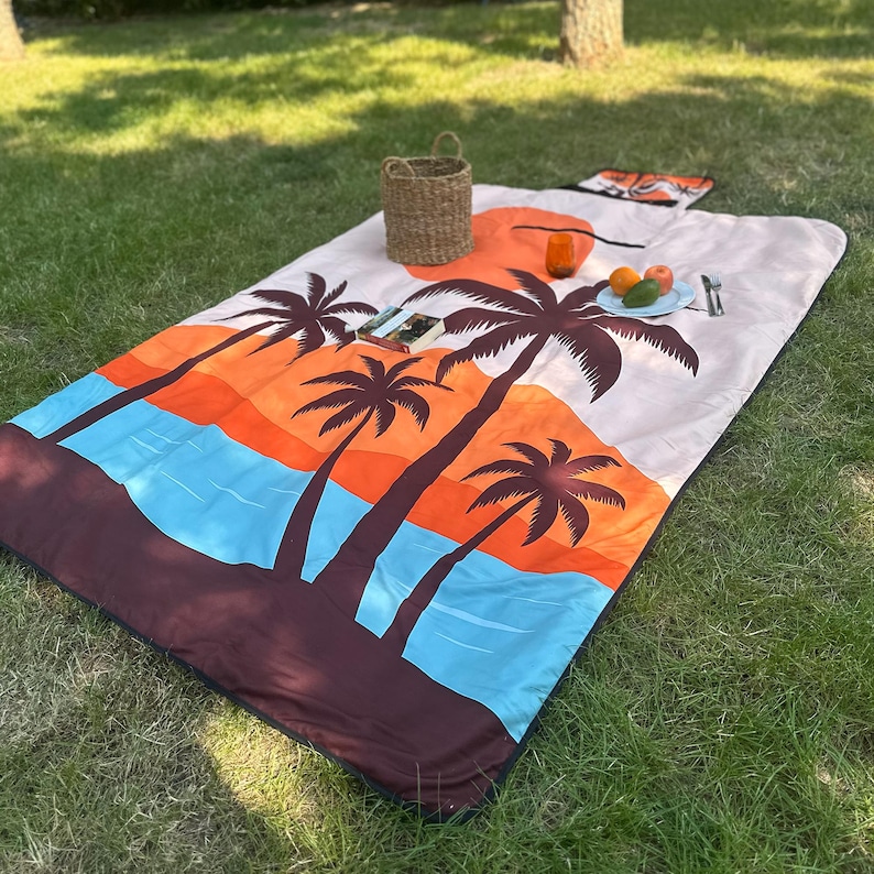 May include: A rectangular picnic blanket with a tropical sunset design featuring palm trees, orange and blue hues. A woven basket, a glass, a plate of fruit, and a book are on the blanket, which is spread on green grass.