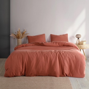 Puede incluir: Un juego de cama de color terracota, que incluye una funda nórdica y dos fundas de almohada, se muestra en una cama. La ropa de cama tiene un aspecto texturizado. La habitación presenta una paleta de colores neutros con una mesa auxiliar tejida y un jarrón decorativo.