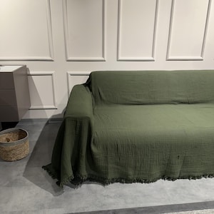 May include: A large, olive green sofa cover made of a textured, crinkled fabric. The cover drapes over the entire sofa, featuring a straight edge along the top and a decorative fringe detail along the bottom hem, providing full coverage.