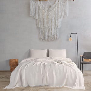 Könnte beinhalten: Ein Schlafzimmer mit einem Bett, das mit einer weißen, strukturierten Decke und Kissen bedeckt ist. Ein gewebter Wandbehang und eine Stehlampe mit einer warmen Glühbirne sind zu sehen. Zwei gewebte Körbe stehen auf dem Boden, und rechts steht ein grauer Stuhl mit Büchern.