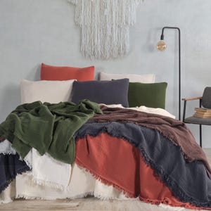 Pode incluir: Uma cama é feita com vários cobertores em tons de verde, castanho, azul marinho e ferrugem. Os cobertores têm uma borda com franjas. Várias almofadas de cor lisa em ferrugem, creme, azul marinho e verde são dispostas na cama. Uma tapeçaria de macramé está acima da cama.