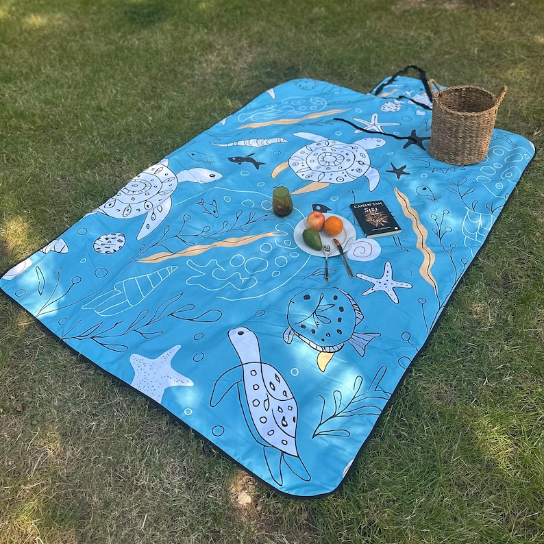 May include: A blue picnic blanket featuring a marine life design with turtles, fish, and starfish. The blanket has a black border and is laid on green grass. A woven basket, a plate of fruit, and a book are on the blanket.