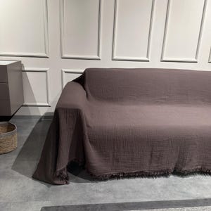 May include: A dark mauve, textured cotton muslin sofa cover draped over a couch. The fabric features a crinkled, multi-layered appearance with a soft, matte finish and a decorative fringe trim along the bottom edge, providing full coverage.
