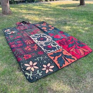 May include: A patterned picnic blanket with a patchwork design. The blanket features squares of red, green, orange, and white, each with a different floral motif. The blanket is spread out on grass. The blanket is rectangular.