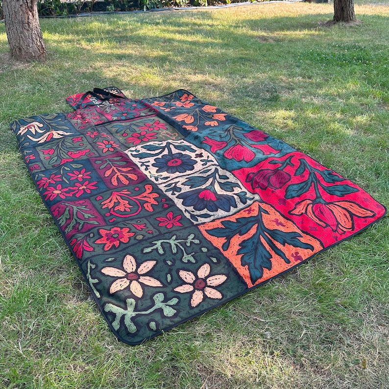 May include: A patterned picnic blanket with a patchwork design. The blanket features squares of red, green, orange, and white, each with a different floral motif. The blanket is spread out on grass. The blanket is rectangular.