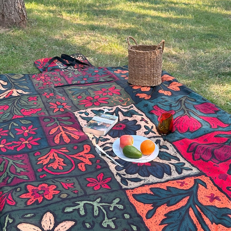 May include: A picnic setting on grass. A large, patterned blanket with floral designs in red, green, and orange is laid out. A woven basket, a book, a plate of fruit, and a small glass are arranged on the blanket.
