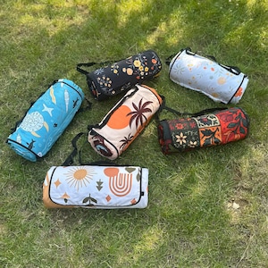 May include: A selection of rolled picnic blankets with carry straps, arranged on green grass. The blankets display diverse patterns, such as floral, celestial, and geometric designs. Colours include blue, orange, black, and white.