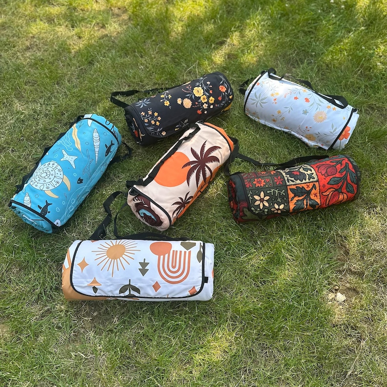 May include: A selection of rolled picnic blankets with carry straps, arranged on green grass. The blankets display diverse patterns, such as floral, celestial, and geometric designs. Colours include blue, orange, black, and white.