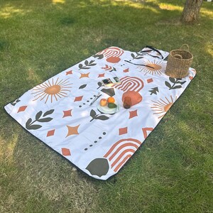 May include: A white picnic blanket with a black border, patterned with orange, brown, and olive green suns, arches, leaves, and diamonds. A woven basket, fruit, and a glass sit on the blanket. The blanket is on grass.