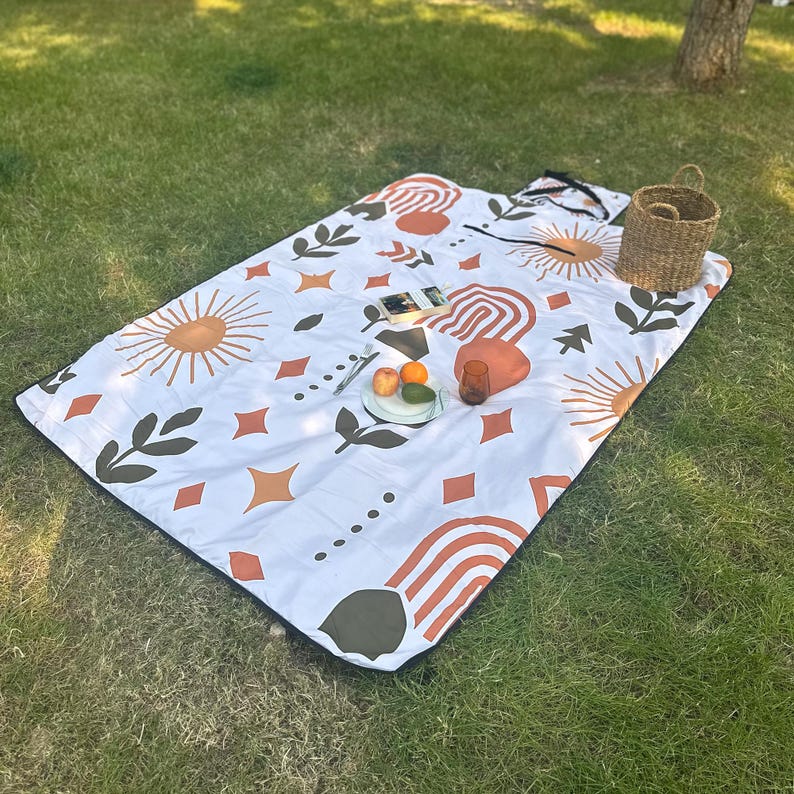 May include: A white picnic blanket with a black border, patterned with orange, brown, and olive green suns, arches, leaves, and diamonds. A woven basket, fruit, and a glass sit on the blanket. The blanket is on grass.
