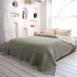 May include: A bed with a sage green waffle-weave blanket and cream-colored pillows. The blanket has a fringed edge and covers the bed. The bed is on a wooden frame, and the room has white walls and a window.