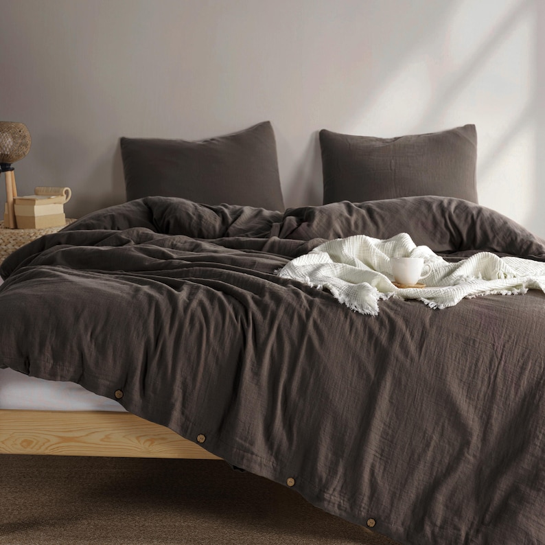 May include: A dark brown bed set with two matching pillowcases. The comforter has wooden buttons along the edge. A white blanket with fringe and a white mug rest on top of the comforter. The bed frame is made of light-colored wood.