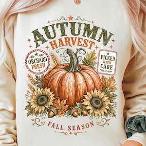 Peut inclure: Un sweat-shirt à col rond couleur crème avec une impression graphique représentant une grosse citrouille orange entourée de tournesols, d'épis de blé et de glands. Le texte indique AUTUMN HARVEST, ORCHARD FRESH, PICKED WITH CARE et FALL SEASON.
