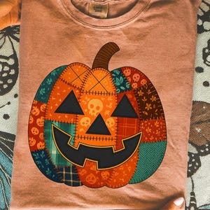 Peut inclure: T-shirt pêche avec un motif citrouille patchwork. La citrouille a une tige marron, des yeux triangulaires noirs et une bouche souriante. La citrouille est composée de divers motifs de tissu, dont des crânes et des motifs floraux.