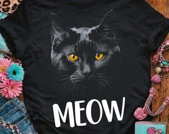 Op de afbeelding: Zwart T-shirt met een afbeelding van een zwarte kattenkop met gele ogen. Het woord "MEOW" staat eronder in het wit gedrukt. Het T-shirt heeft een ronde hals.