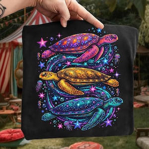 May include: Black tote bag featuring a vibrant illustration of three colorful sea turtles. The turtles are surrounded by swirling patterns, stars, and jellyfish in shades of purple, blue, and gold. The design is held by a hand.