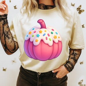 Peut inclure: T-shirt crème avec une grande citrouille rose de style dessin animé. La citrouille est ornée d'un glaçage blanc, de vermicelles colorés et d'une tige rose. Le t-shirt est un vêtement décontracté, idéal pour un usage quotidien.