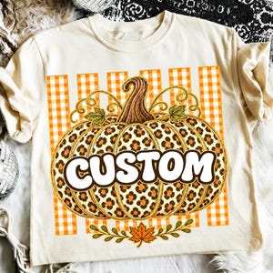 Peut inclure: Un t-shirt couleur crème arborant un motif central de citrouille avec un imprimé léopard. La citrouille est placée sur un fond à rayures vichy orange et blanc et comporte le mot CUSTOM écrit en lettres blanches grasses.