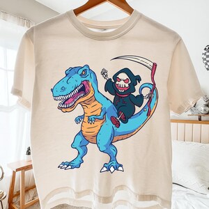 Pode incluir: Camiseta creme com uma ilustração de desenho animado. Apresenta um Tyrannosaurus Rex azul com detalhes em laranja, montado por uma figura da Morte. A Morte, em um manto preto com capuz, segura uma foice. O design está centrado na camiseta.
