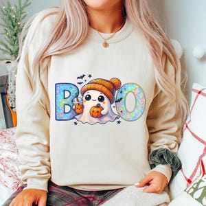 Peut inclure: Un sweat-shirt crème avec un motif sur le thème d'Halloween. Le mot "BOO" est écrit avec un personnage fantôme mignon portant un bonnet orange, tenant une citrouille et une boisson. Des chauves-souris et des étoiles complètent le design festif.