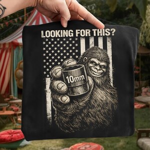 Peut inclure: T-shirt noir avec un motif de Bigfoot souriant tenant une clé à douille de 10 mm. Le design comprend un drapeau américain et le texte "LOOKING FOR THIS ?" en blanc. L'image est une représentation humoristique d'un outil de mécanicien.