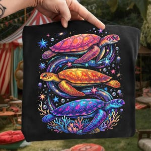 May include: Black tote bag featuring a vibrant illustration of three colorful sea turtles. The turtles are depicted in shades of pink, orange, and blue, surrounded by stars, galaxies, and aquatic elements. The design is detailed and eye-catching.