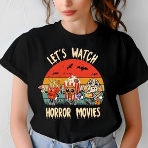 Peut inclure: T-shirt noir avec un motif de personnages de dessins animés : frites, pop-corn et un personnage masqué. Le texte "Let's Watch Horror Movies" est au-dessus des personnages, avec un coucher de soleil et des chauves-souris en arrière-plan.