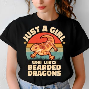 Peut inclure: T-shirt noir avec un motif de dragon barbu dans un cercle de coucher de soleil. Le texte dit "JUST A GIRL WHO LOVES BEARDED DRAGONS" en blanc et jaune. Le design est de style rétro.