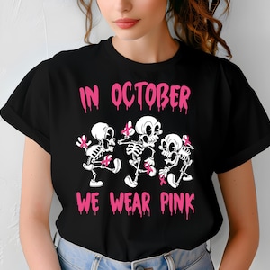 Peut inclure: T-shirt noir avec l'inscription rose "IN OCTOBER WE WEAR PINK" au-dessus de trois squelettes dansant avec des rubans roses. Les squelettes ont des crânes et des os blancs. Le t-shirt est porté par une personne.