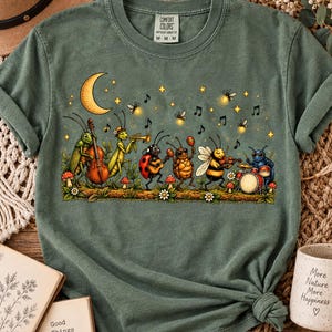 Könnte beinhalten: Ein salbeigrünes T-Shirt mit einer Grafik von Insekten, die Musikinstrumente spielen, darunter eine Grille mit Cello, eine Heuschrecke mit Trompete, ein Marienkäfer mit Gitarre, ein Käfer mit Maracas, eine Biene mit Geige und ein Käfer am Schlagzeug.