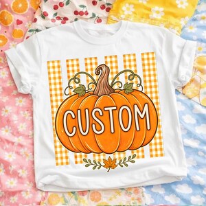 Peut inclure: Un t-shirt blanc avec un grand motif de citrouille orange centré sur un fond à rayures vichy jaune et blanc. Le mot CUSTOM est imprimé en lettres capitales blanches sur la citrouille, qui est ornée de vrilles vertes.